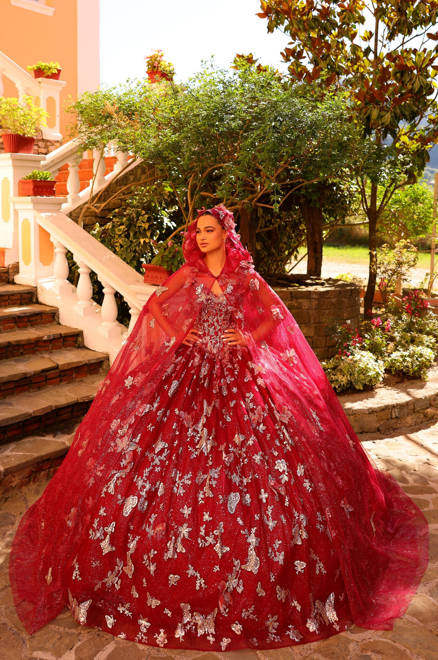 A woman in a red butterfly-embellished ballgown poses on stone steps with lush garden backdrop.