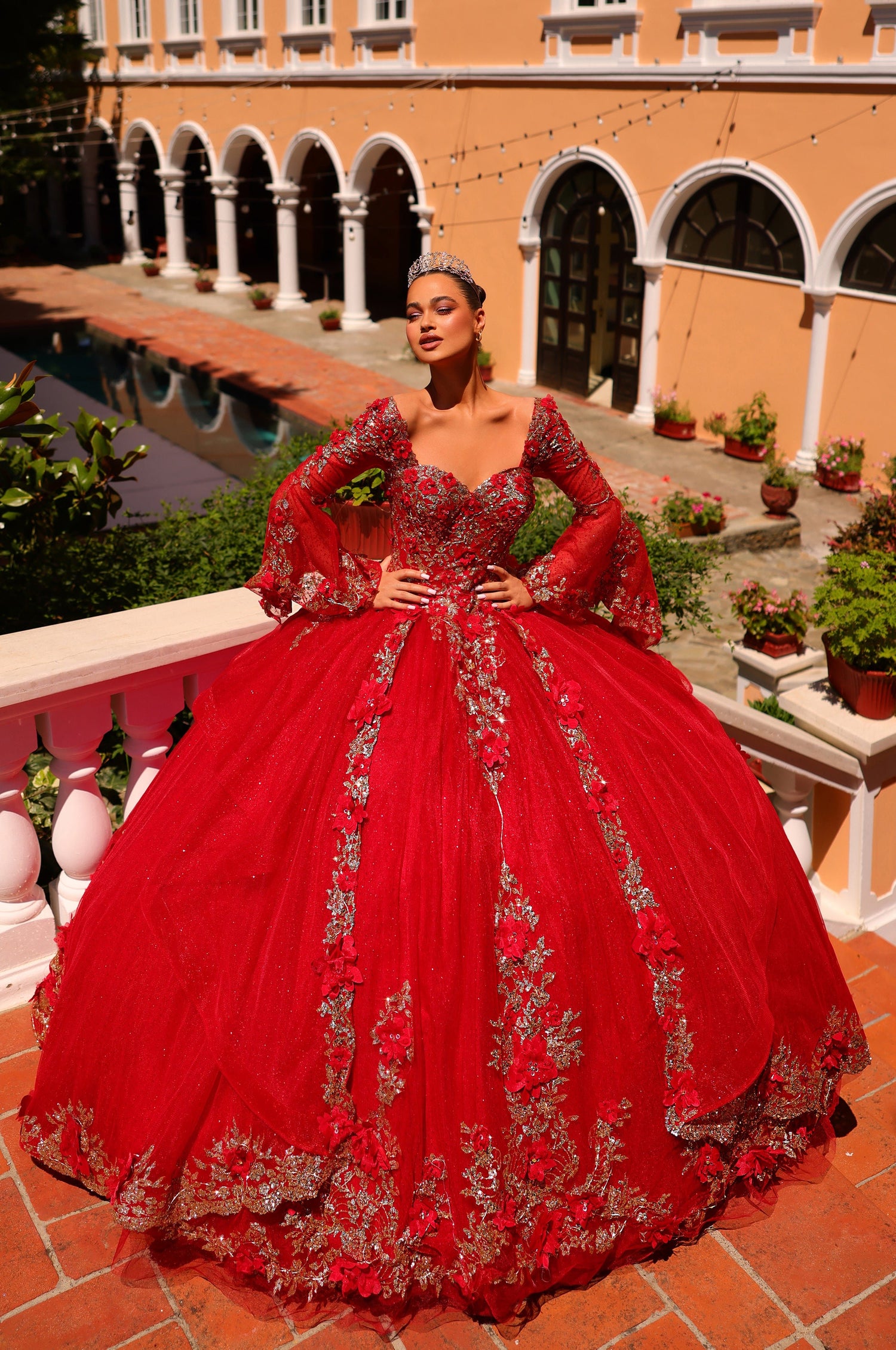 A woman wears a vibrant red ballgown with silver embellishments, standing in an arched courtyard.