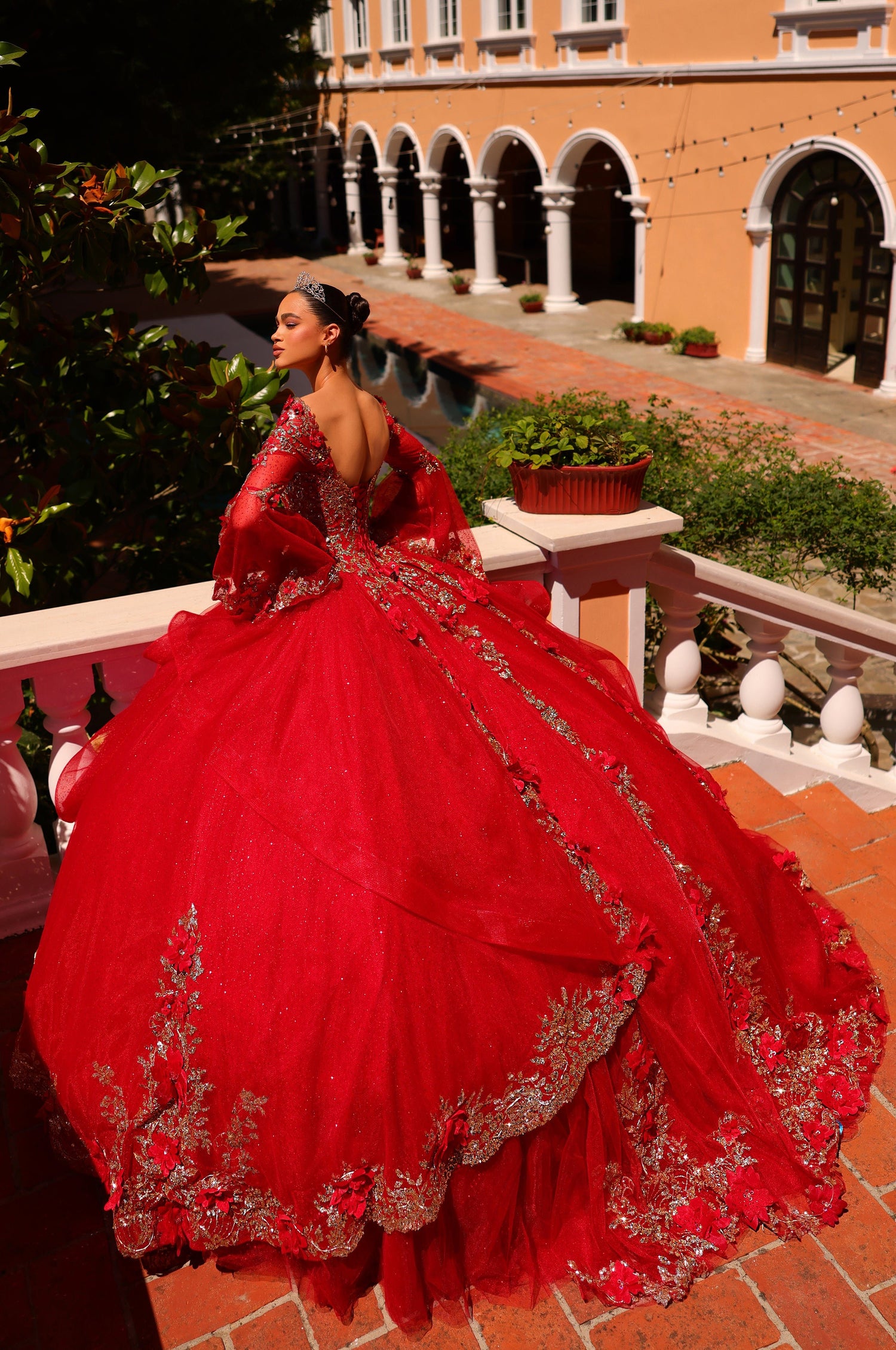 A person in a vibrant red ballgown with silver embroidery poses on a tiled terrace with arched architecture.