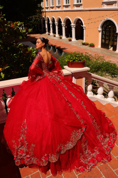 A person in a vibrant red ballgown with silver embroidery poses on a tiled terrace with arched architecture.