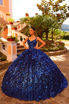 A woman wears a voluminous royal blue floral lace ballgown in a sunlit garden with stone steps.