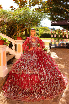 A model wears an elaborate Wine/Gold quinceañera ball gown with intricate floral embroidery in a garden setting.