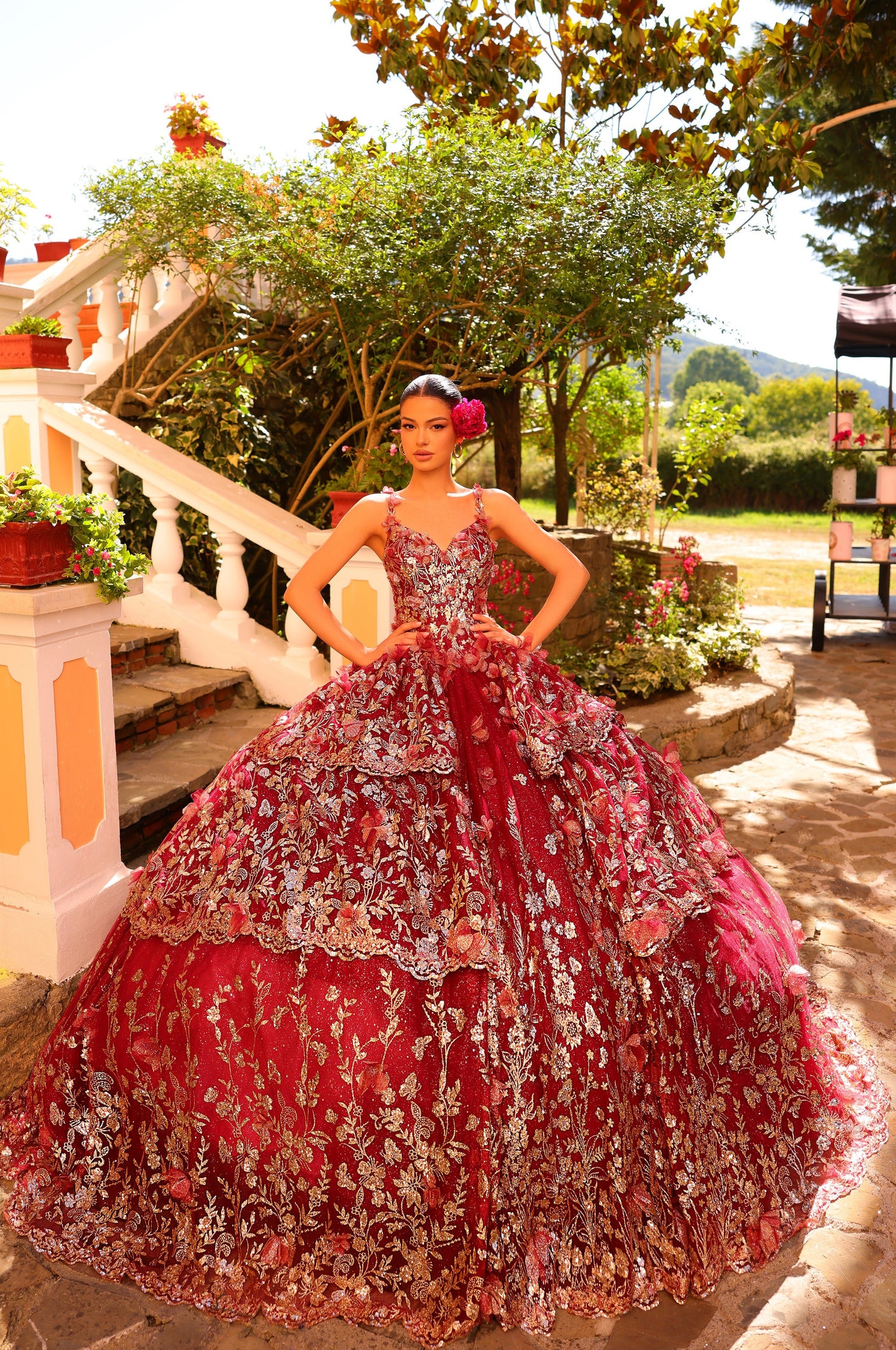 A woman in a red floral embroidered ballgown stands on a stone patio with gardens and trees in the background.