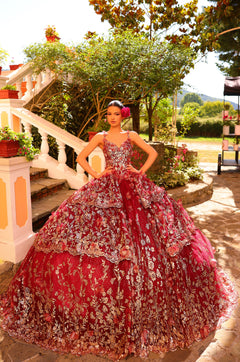 A woman in a red floral embroidered ballgown stands on a stone patio with gardens and trees in the background.