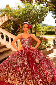 A woman wears an ornate red quinceañera ball gown with floral embroidery in a garden setting.