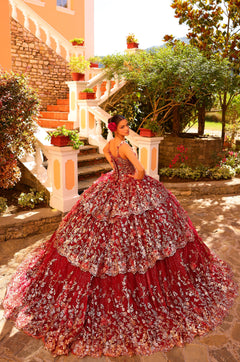 A woman in a red and silver floral quinceañera gown poses on stone steps with lush garden surroundings.