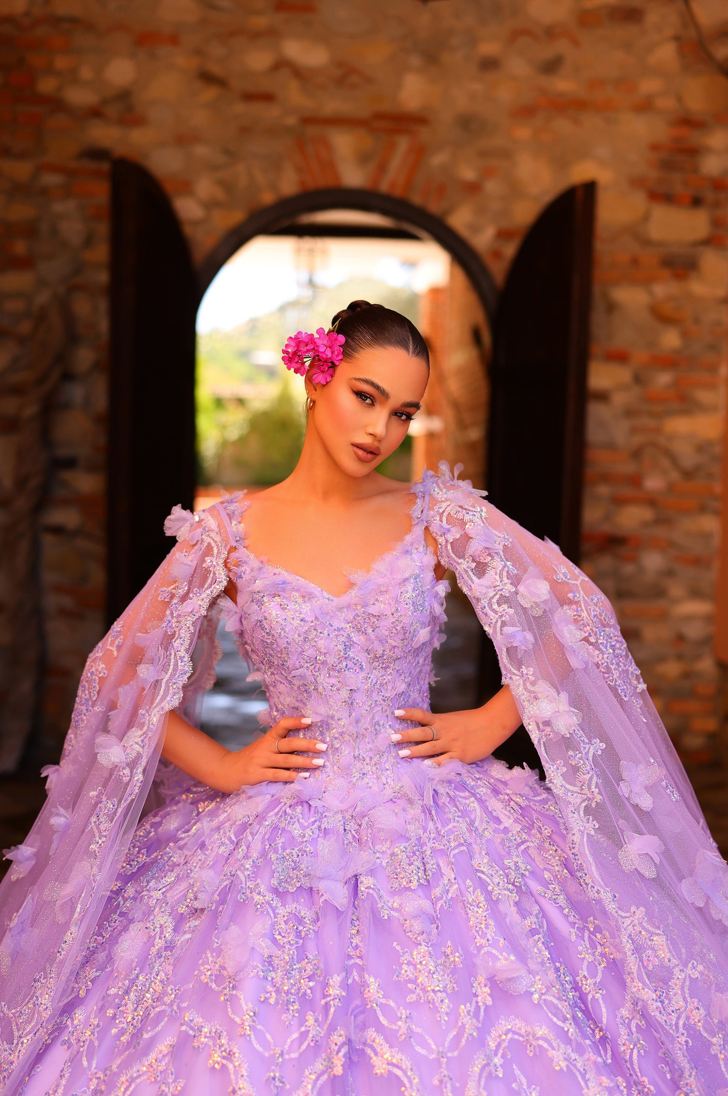 A woman in a lavender lace ball gown poses in an arched stone window with a bright pink flower in her hair.