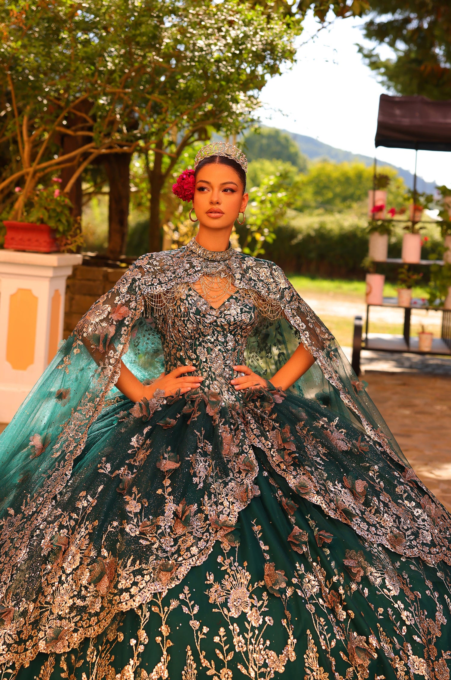 A model wears an emerald green quinceañera ball gown with intricate gold floral embroidery and a matching cape.