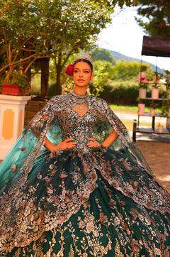 A model wears an emerald green quinceañera ball gown with intricate gold floral embroidery and a matching cape.