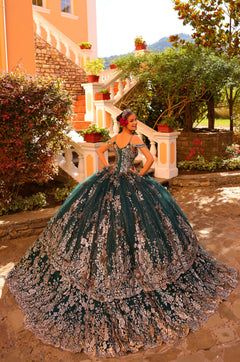 A person in an emerald green, floral-embroidered quinceañera ball gown poses on stone steps with gardens behind.