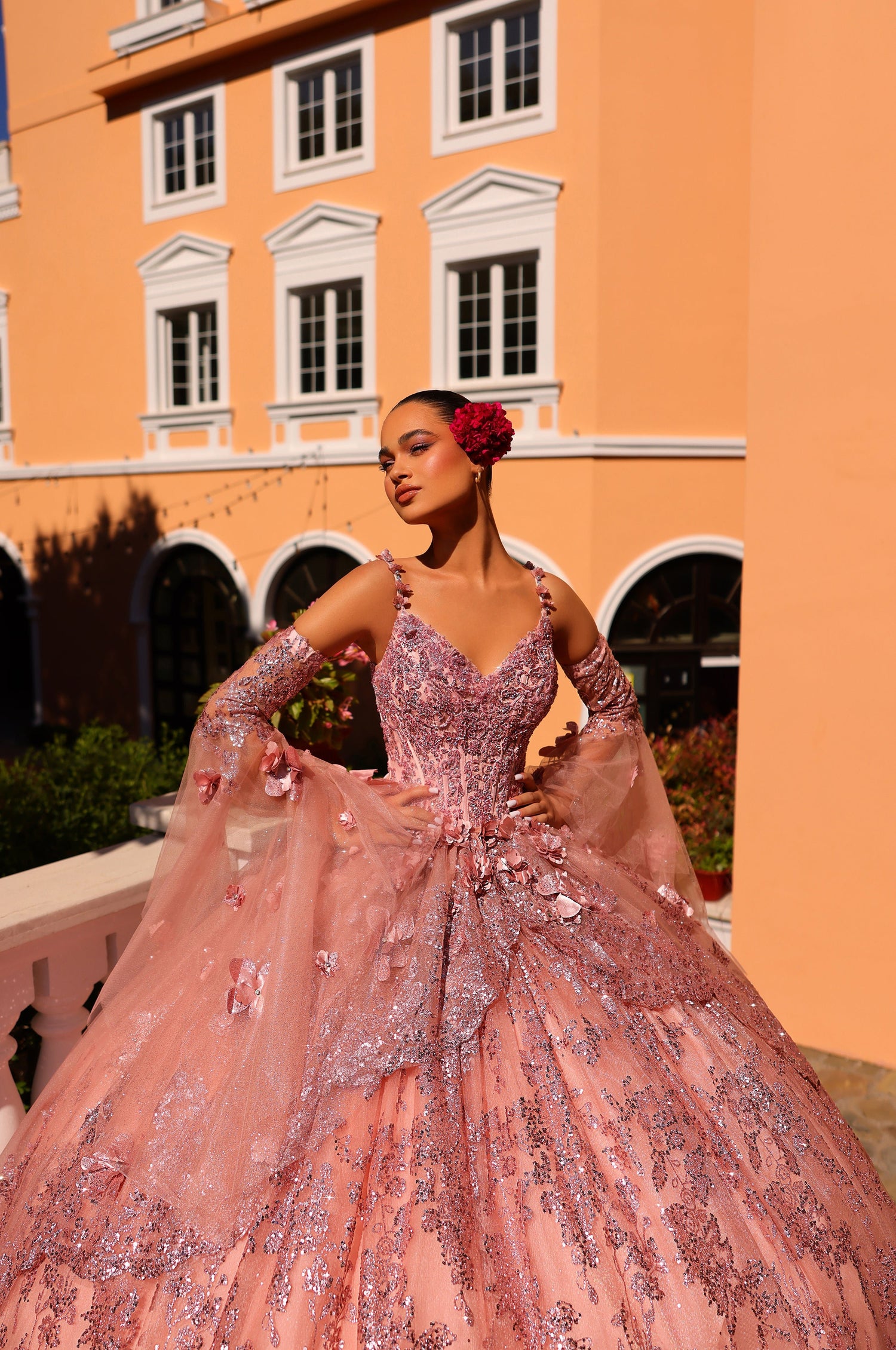 A model wears a sparkling pink ball gown with 3D floral details, posing against an orange building.