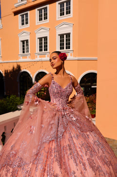 A model wears a sparkling pink ball gown with 3D floral details, posing against an orange building.