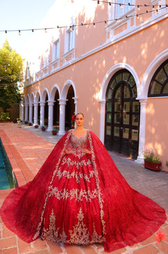 A woman wears an ornate red ballgown with silver embroidery, standing before a columned building with string lights.
