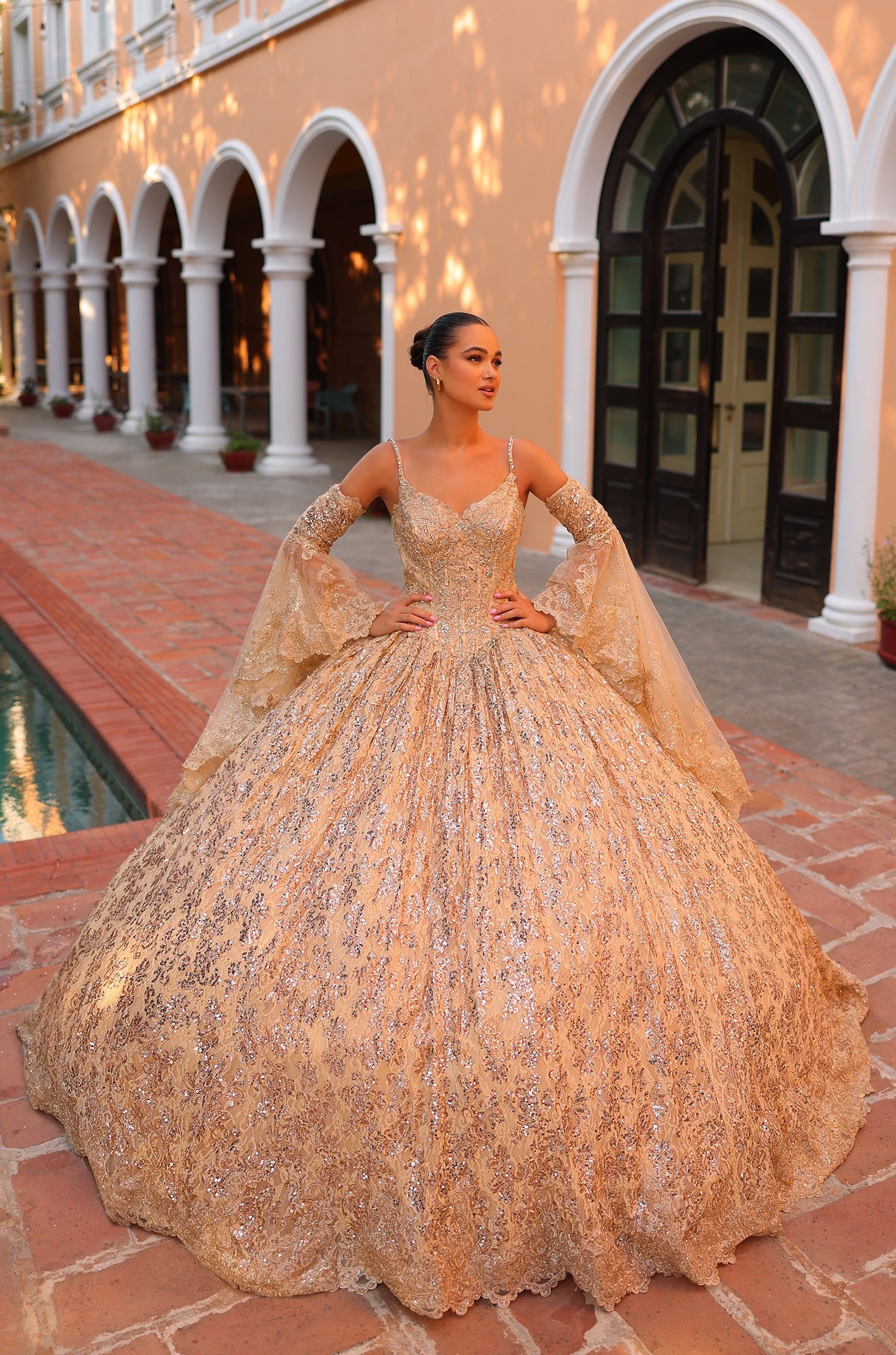 A woman wears a sparkling Gold ball gown with intricate lace and bell sleeves, standing in an arched architectural setting.