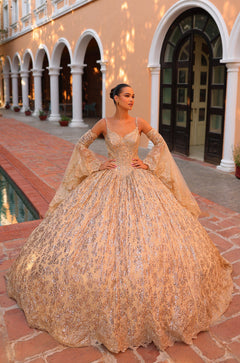 A woman wears a sparkling Gold ball gown with intricate lace and bell sleeves, standing in an arched architectural setting.