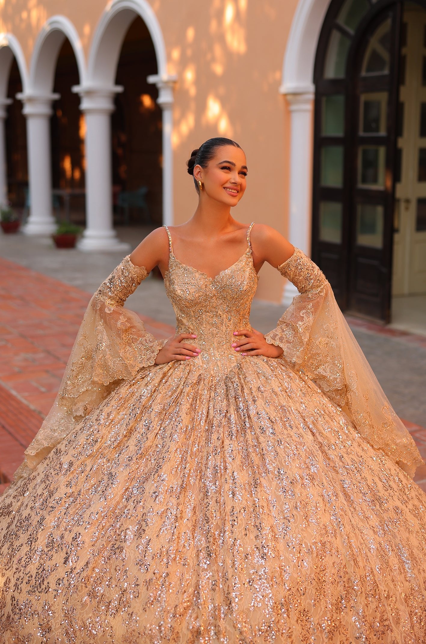 A woman in a glittering gold ballgown with lace sleeves poses confidently in an arched architectural setting.