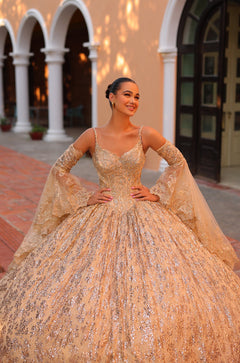 A woman in a glittering gold ballgown with lace sleeves poses confidently in an arched architectural setting.