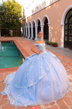 A young woman wears a light blue ballgown with floral embroidery, posing by a pool near a Spanish-style building.