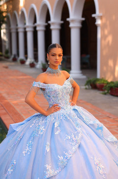 A woman in a blue ballgown with floral embellishments stands confidently in an arched columned courtyard.