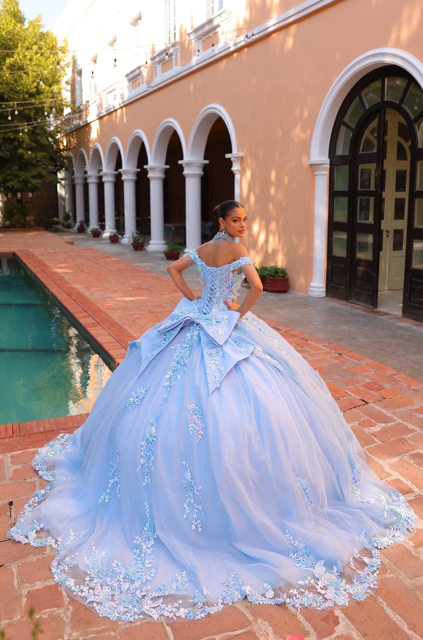 A young woman in a light blue, floral-embroidered ballgown poses by a pool near columned architecture.