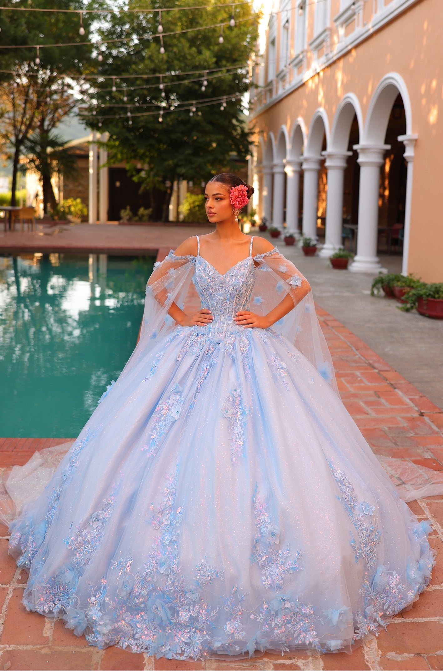 A young woman wears an elaborate Light Blue ball gown with floral details, standing by a pool in an arched courtyard.