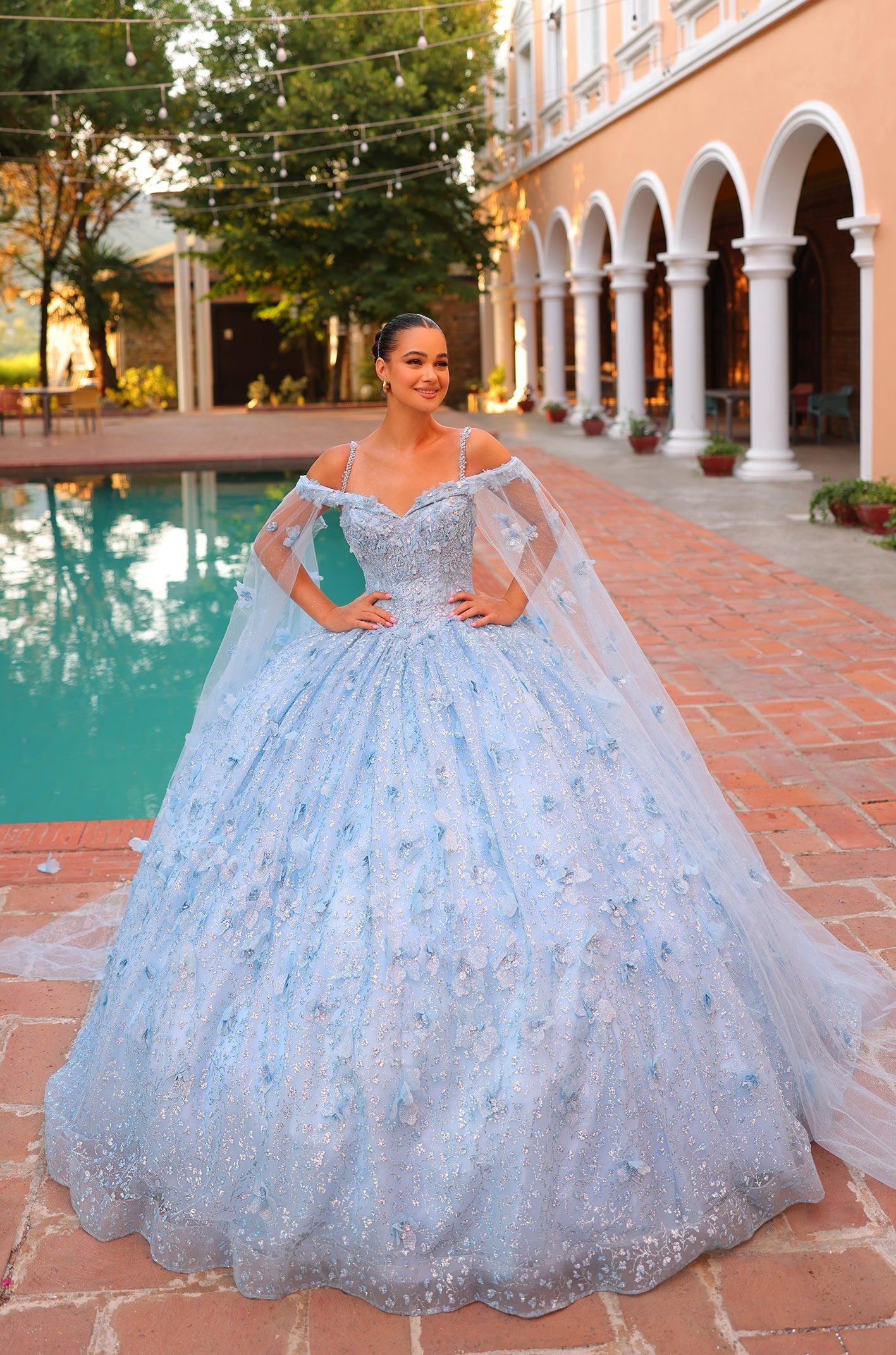 A young woman wears a light blue ball gown with sparkling details, standing by a pool in an elegant courtyard.