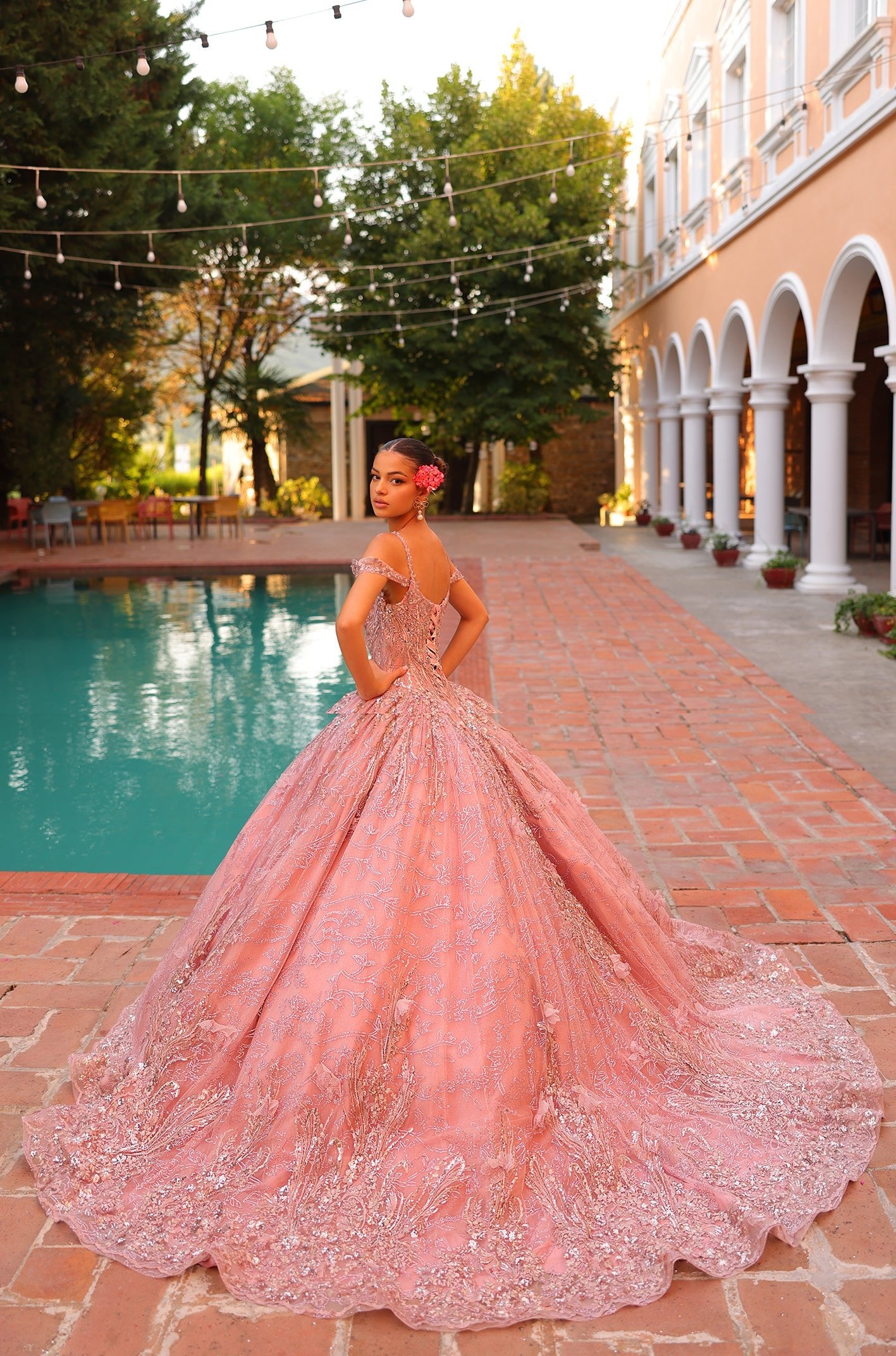 A young woman in a pink ball gown poses by a pool with decorative string lights and arched architecture.
