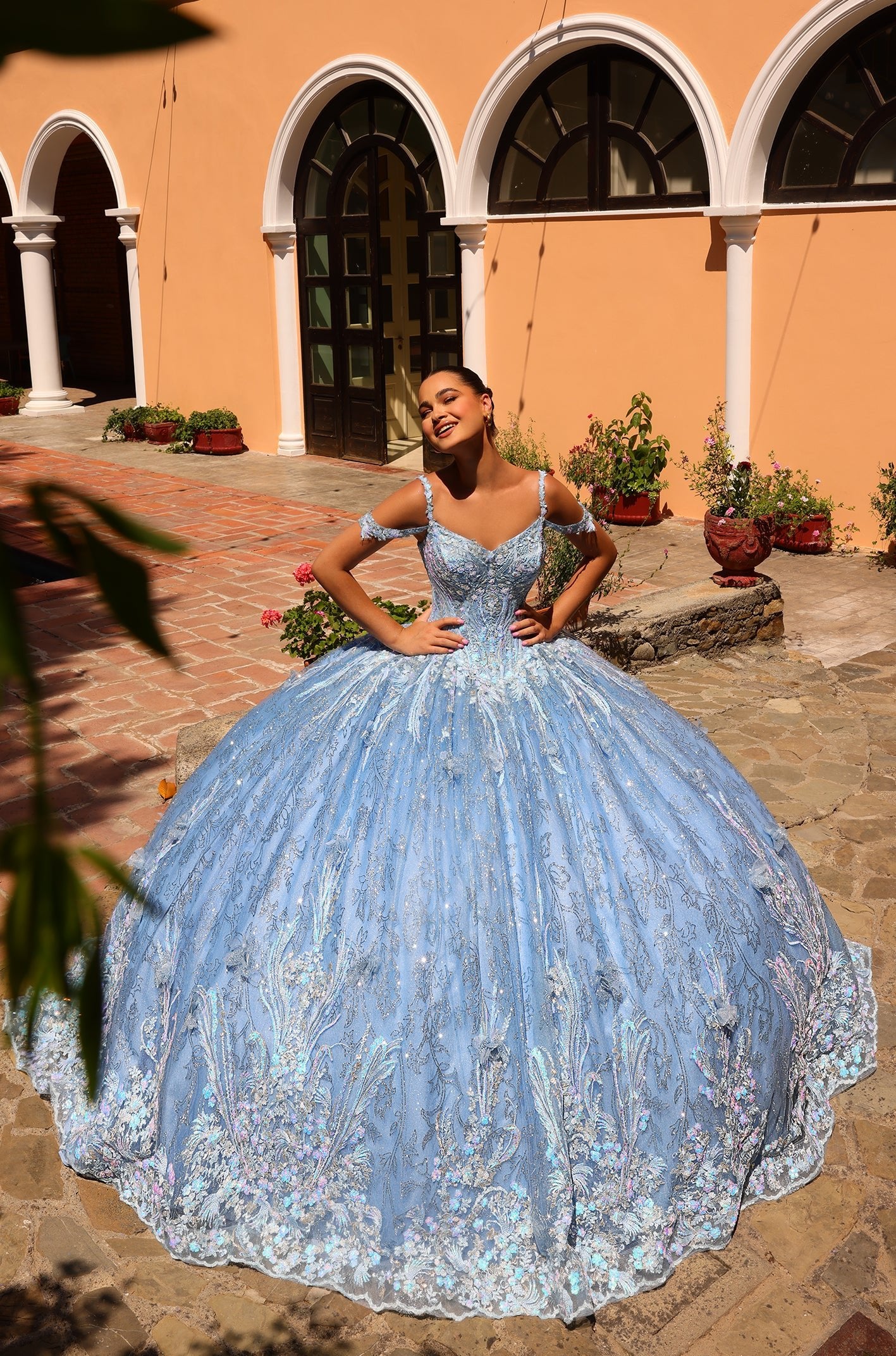 A smiling quinceañera poses in a sparkly Bahama Blue ballgown with an expansive skirt against a terracotta-walled backdrop.