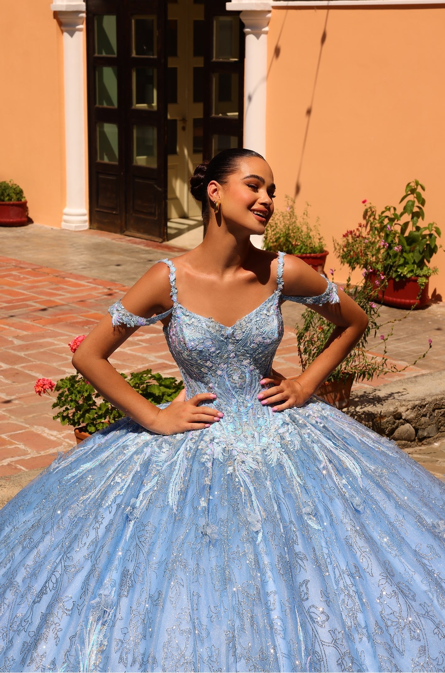A young woman wears a blue sparkly ball gown, posing confidently on a brick patio with potted plants.