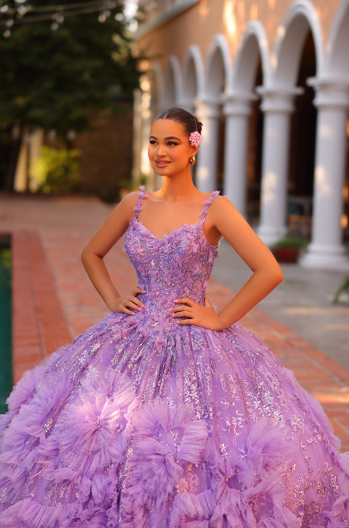 A young woman wears a lavender ball gown with floral embellishments, posing confidently on a brick pathway.