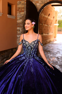 A woman in a deep blue ball gown with off-shoulder straps stands smiling in a stone archway.