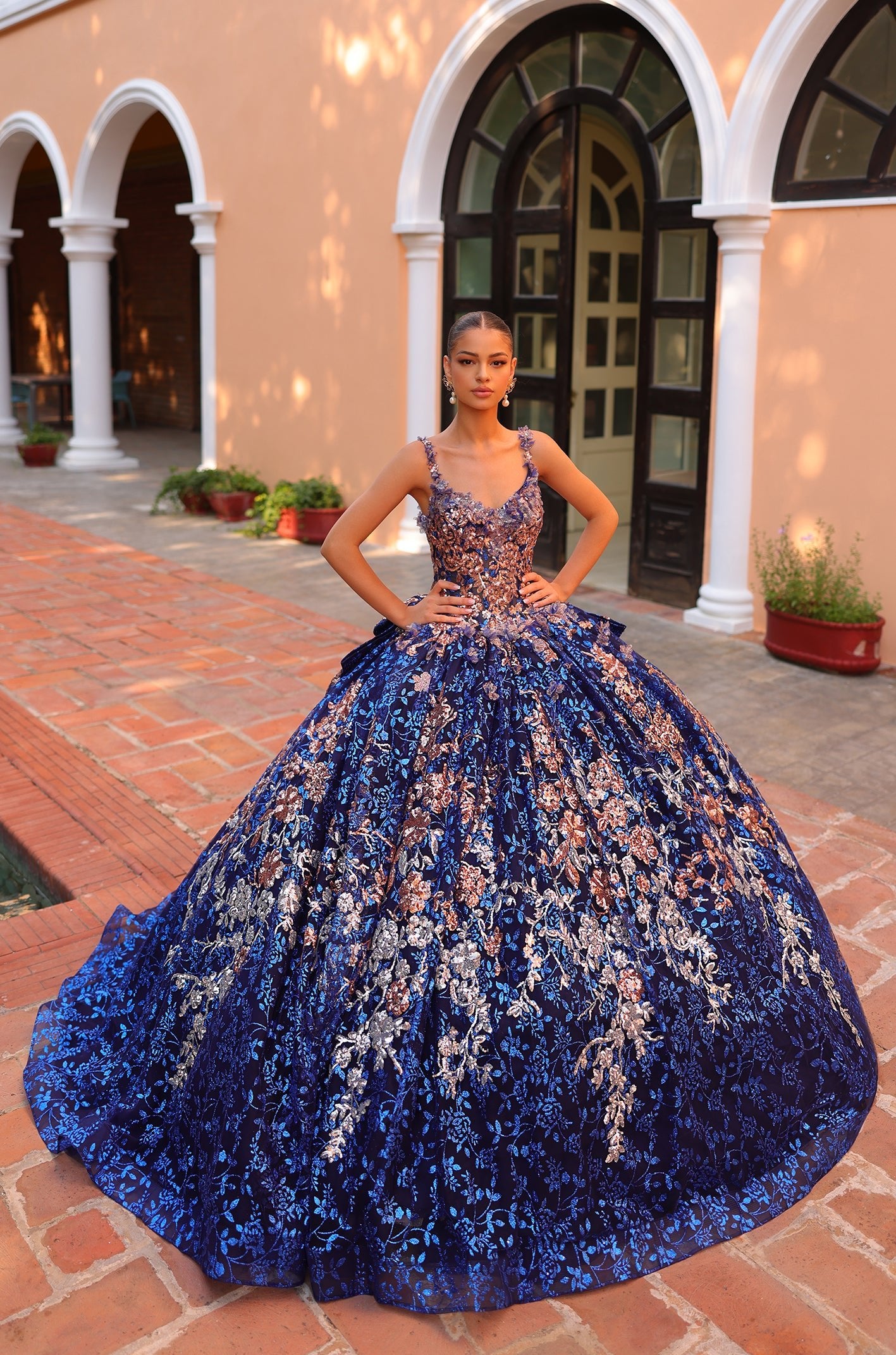 A model wears a Royal Blue/Multi floral ball gown with intricate embellishments on brick-lined architectural grounds.