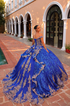 A woman wears an ornate royal blue ball gown with gold lace, posing in an arched courtyard with brick floors.