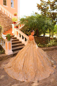 A golden quinceañera gown with intricate embellishments cascades dramatically on stone steps amid lush greenery.