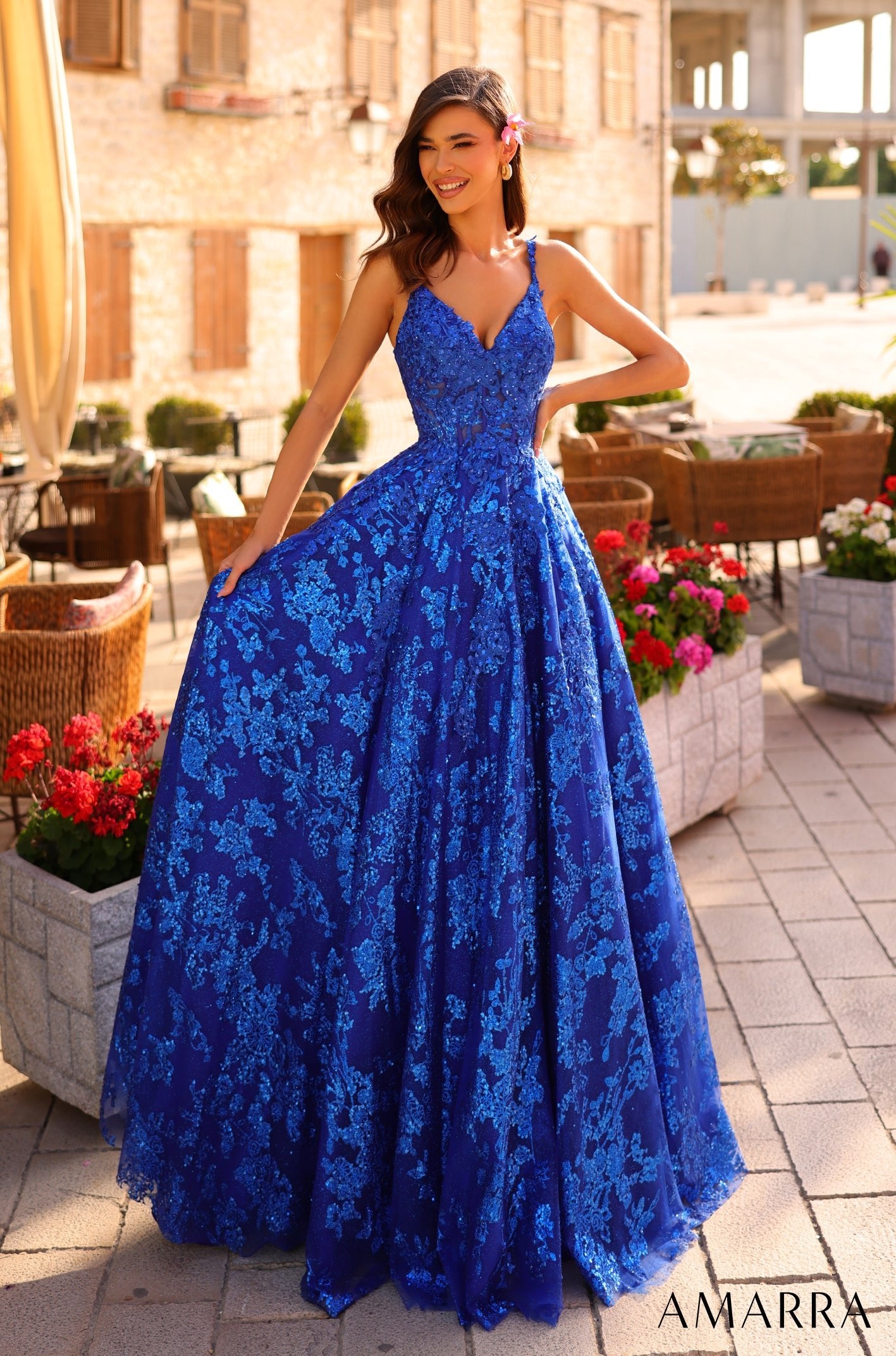 A woman wears a shimmering Royal Blue lace ball gown, standing amid flowers and stone planters.