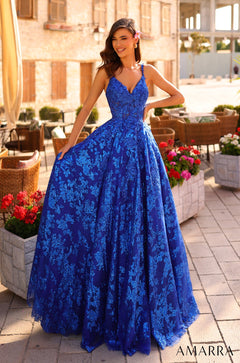 A woman wears a shimmering Royal Blue lace ball gown, standing amid flowers and stone planters.