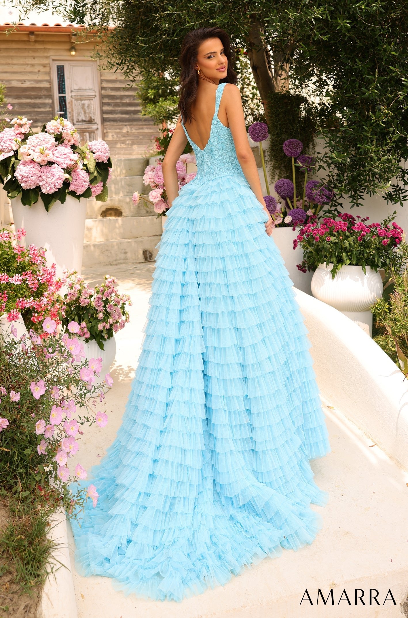 A woman in a blue tiered tulle gown stands in a garden with pink and purple flowers.