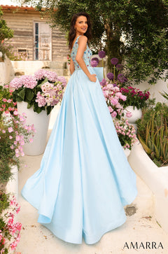 A woman wears a light blue ball gown with floral embellishments, standing in a garden with pink and purple flowers.