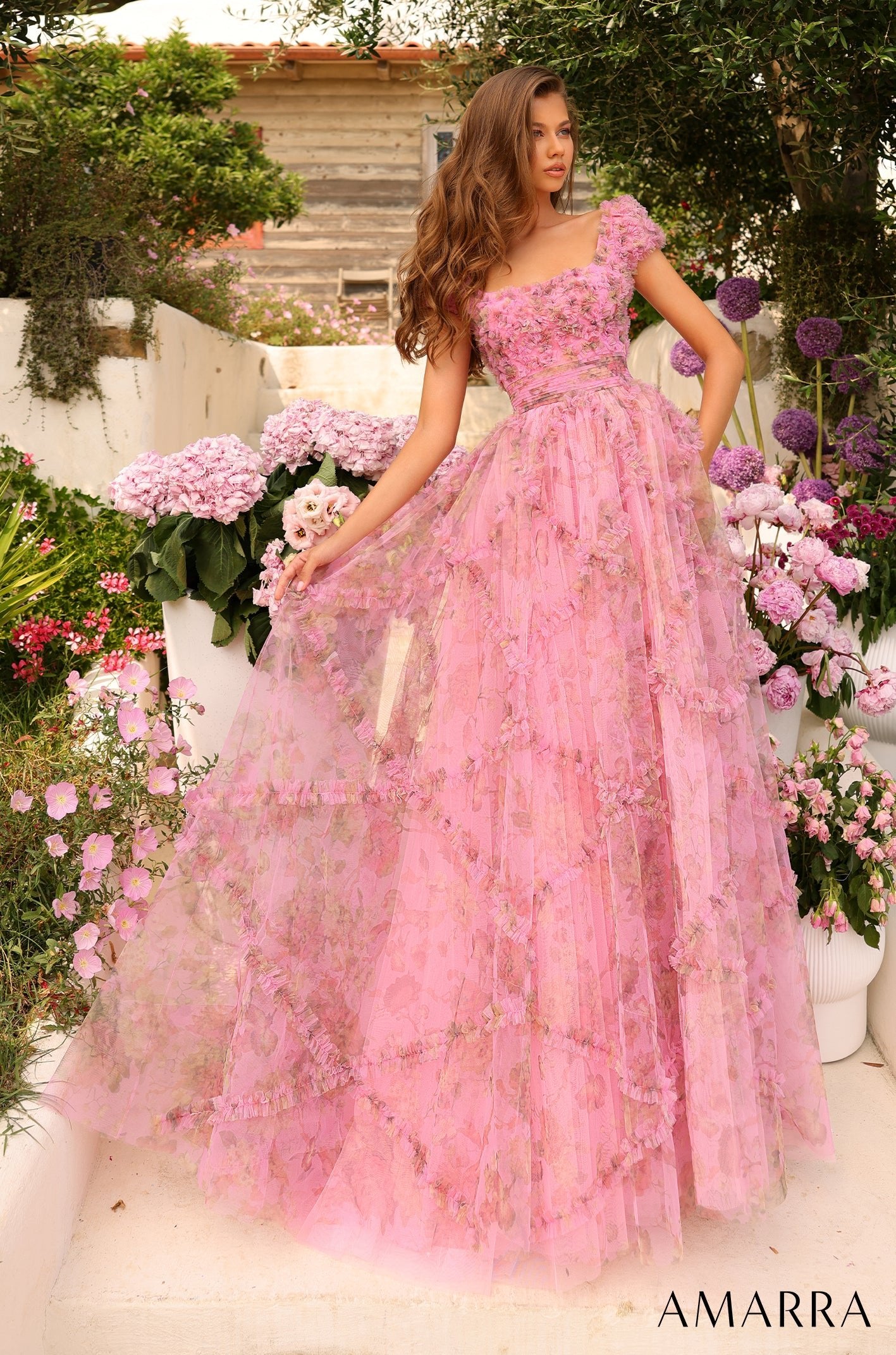 A model wears a pink floral-appliqué ball gown amid lush garden flowers and greenery.