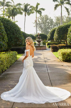 A bride in a white lace mermaid wedding dress stands on a path with palm trees and a fountain in the background.