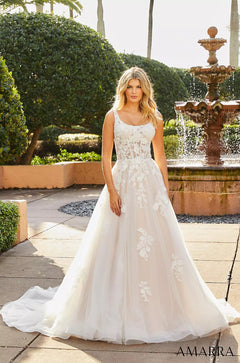 A blonde woman wears a white lace ballgown with floral appliqués, standing near a tiered fountain in a garden.