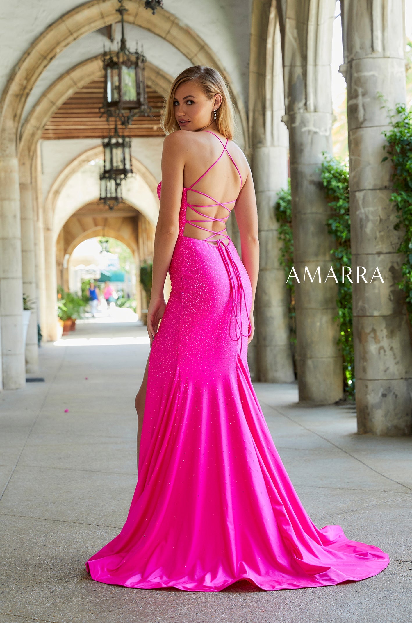 A model wears a bright pink floor-length gown with a strappy back and side slit in an arched stone corridor.