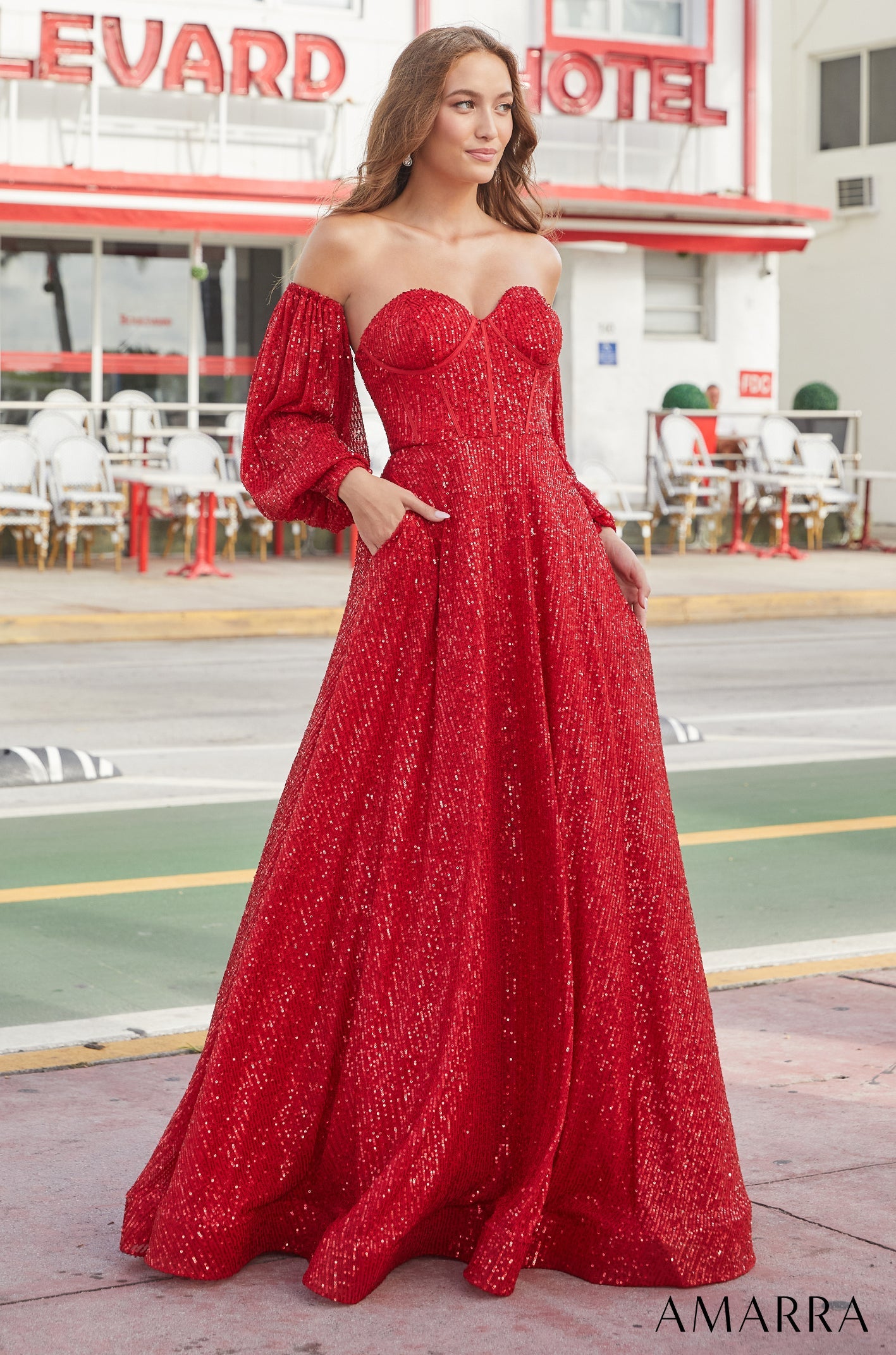 A woman wears a sparkling red off-shoulder ball gown with puffy sleeves in front of a hotel.