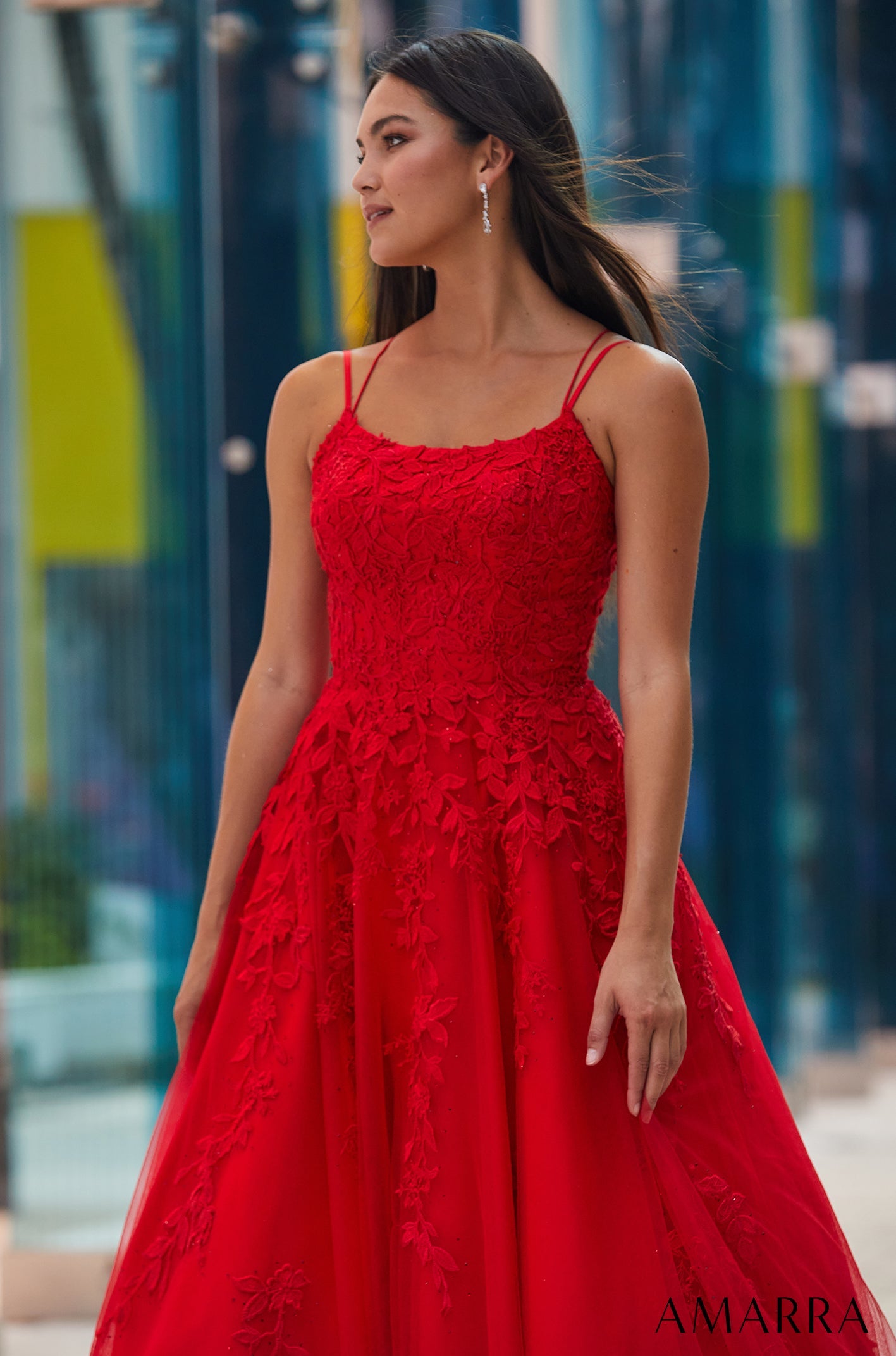 A woman in a vibrant red lace ball gown stands confidently with windblown hair against an urban background.