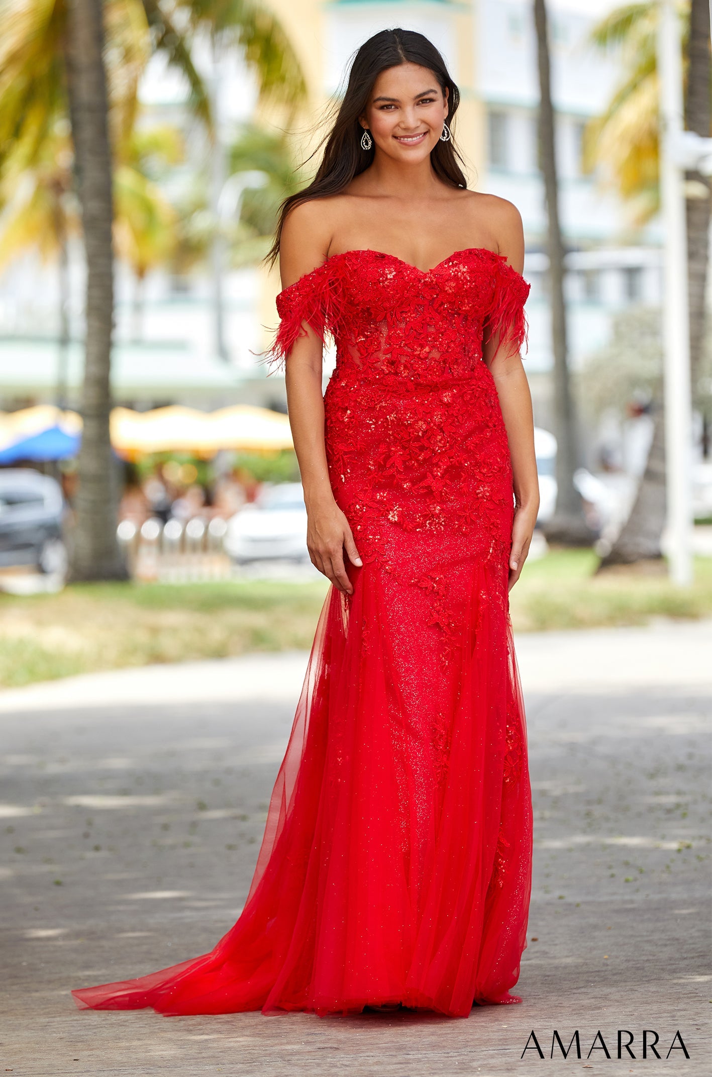 A woman wears a bright red sequined off-shoulder feathered gown against a palm tree backdrop.