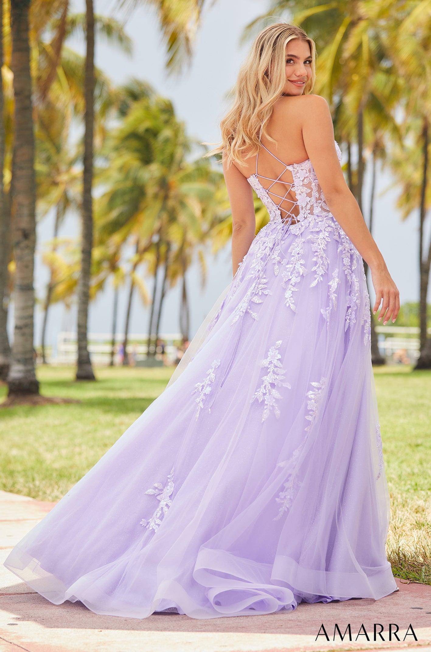 A woman in a lavender ballgown with floral appliqués poses among palm trees, showcasing a lace-up back.