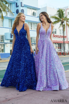 Two women wear sparkling blue and purple floor-length gowns on a palm-lined street with blue buildings.