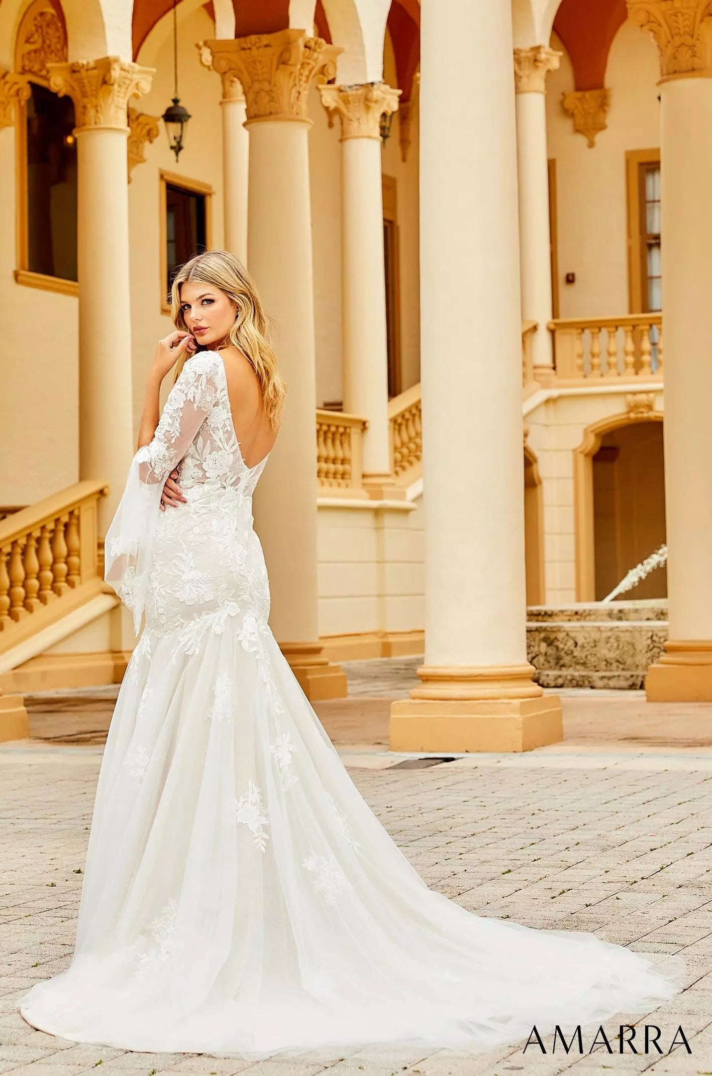 A blonde bride in a white lace mermaid wedding dress poses elegantly among ornate columns.