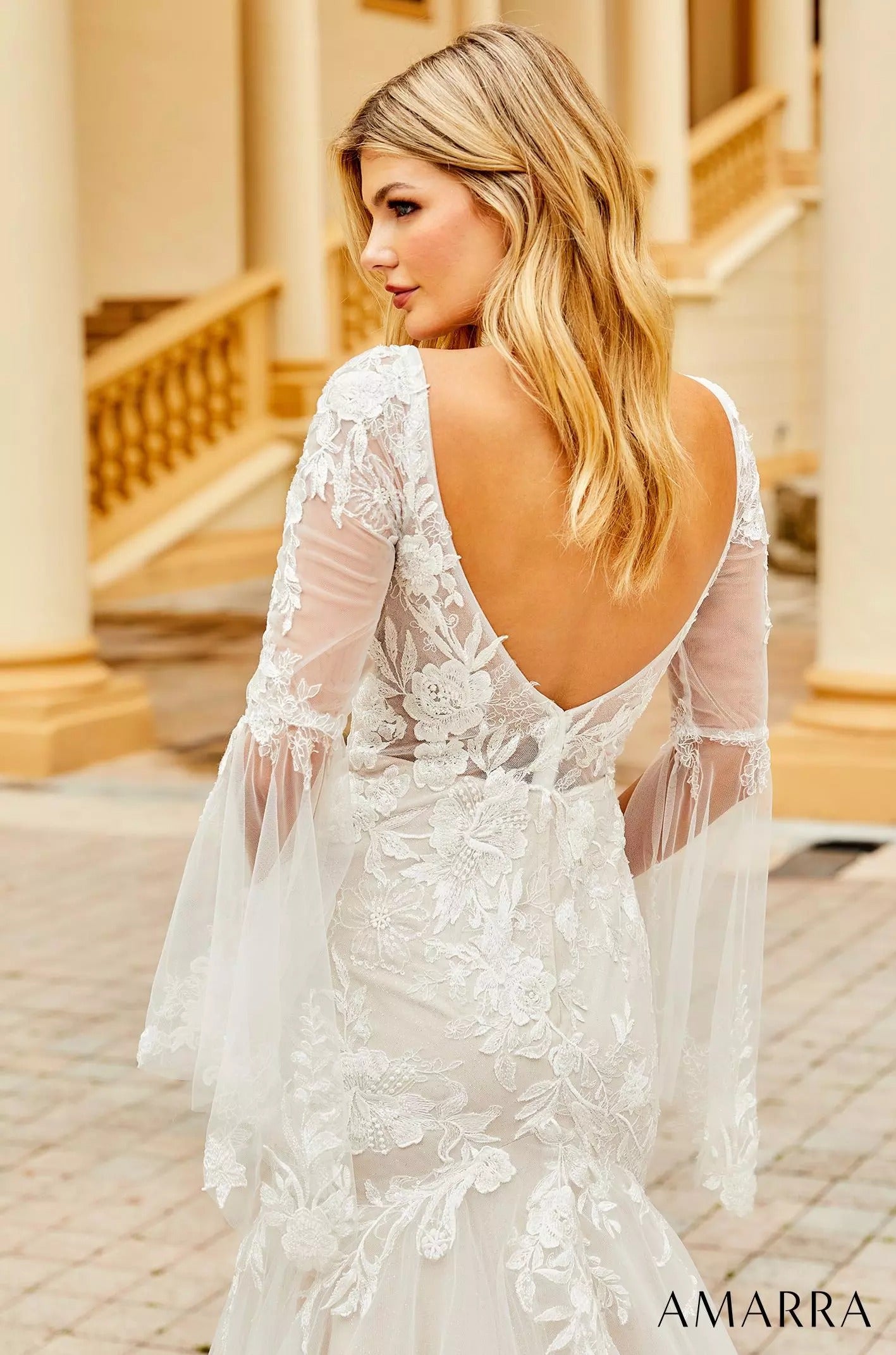 A woman in a white lace wedding dress with sheer sleeves turns, showcasing an intricate floral back design.