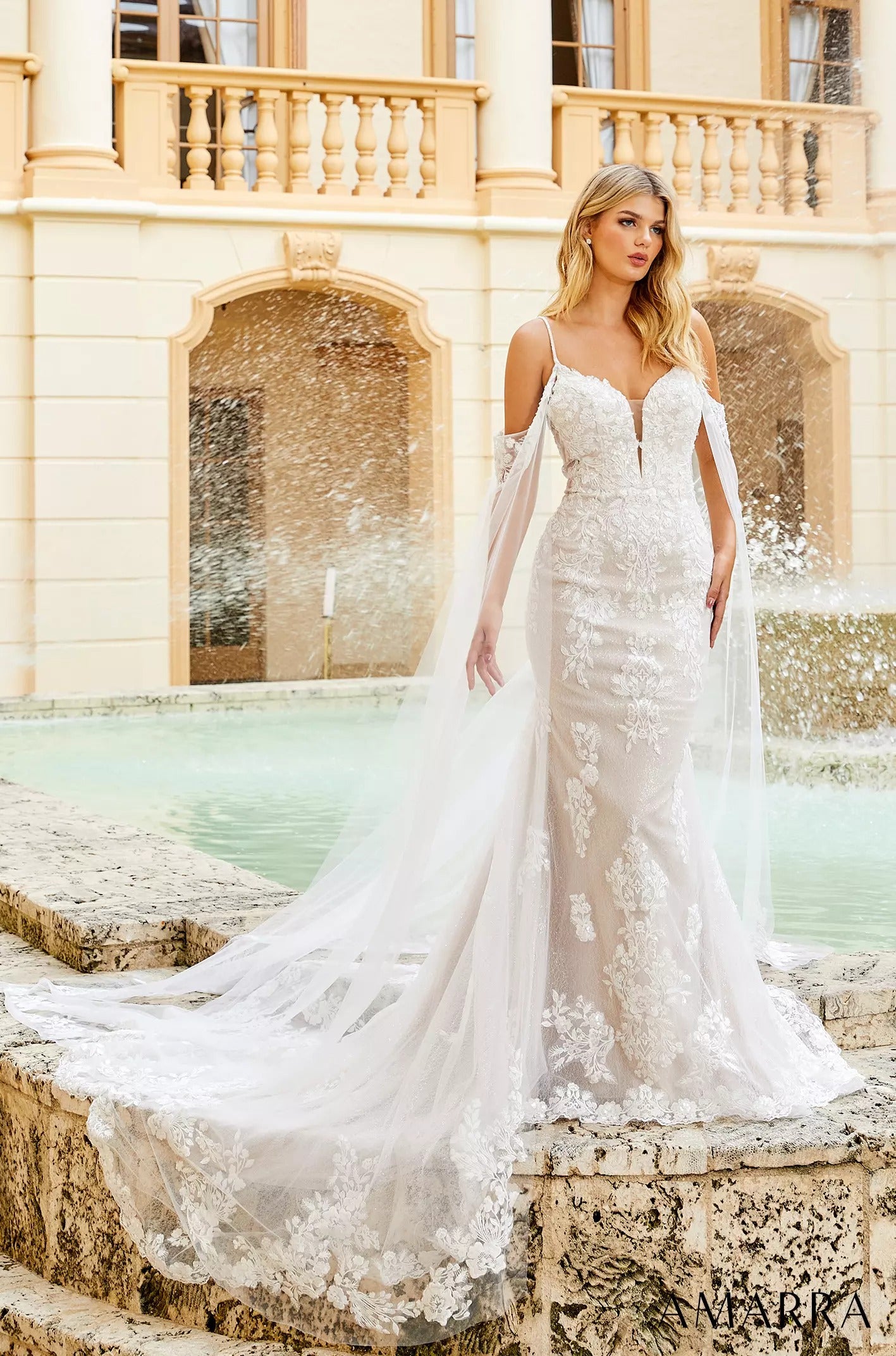 A bride in an elegant white lace mermaid wedding dress with long train stands by a fountain near a classical building.
